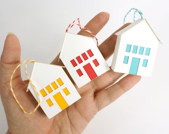 May include: Three white paper house ornaments with colorful accents are held in a hand. Each house has a different colored door and window design: yellow, red, and blue. The ornaments are strung with twine.
