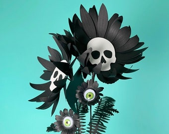 May include: A unique bouquet of paper flowers with a spooky theme. The arrangement features black petals, white skulls, and flowers with green-eyed centers. Ferns and mushrooms add to the gothic aesthetic. The bouquet is held against a teal background.