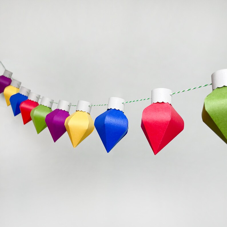 May include: A string of colorful paper ornaments in the shape of Christmas lights. The ornaments are red, yellow, blue, purple, and green. The ornaments are strung on a white string with green accents.