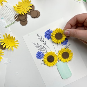 May include: A paper craft bouquet of sunflowers and blue flowers in a light green mason jar. The flowers are made of paper and are arranged in a vase. The background is white.