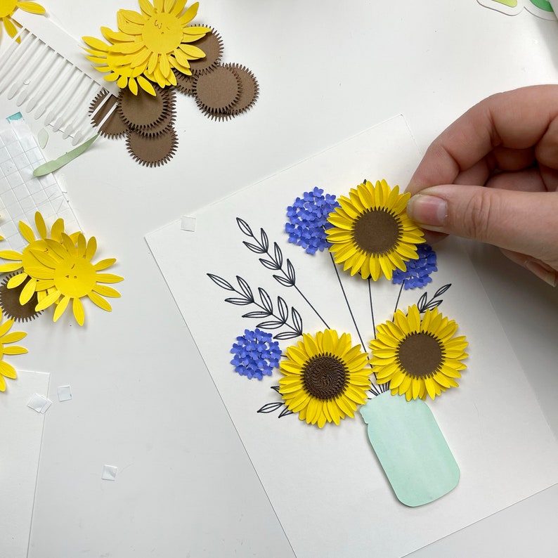 May include: A paper craft bouquet of sunflowers and blue flowers in a light green mason jar. The flowers are made of paper and are arranged in a vase. The background is white.