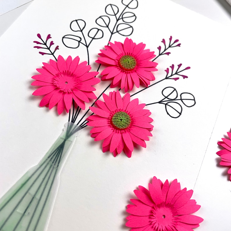 May include: A paper flower arrangement featuring bright pink gerbera daisies with green centres, black stems, and sprigs of purple accents. The flowers are arranged in a clear vase with green accents, set against a white background.