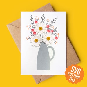May include: A white card featuring a grey vase with a bouquet of white daisies and pink accents. The vase has a handle. The card includes the text "SVG CUTTING FILE" in a circle.