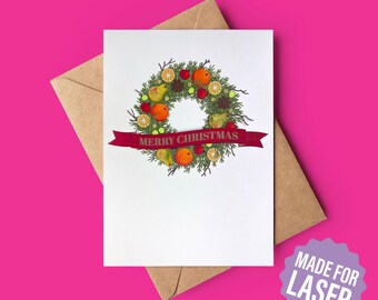 May include: A white greeting card with a festive wreath design. The wreath features oranges, pears, and berries, with a red banner that reads "MERRY CHRISTMAS." The card is on a brown envelope, set against a bright pink background.
