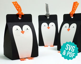 May include: Three black and white penguin-shaped gift boxes with orange beaks and feet. The boxes have a hole at the top for a ribbon. The boxes are labeled "SVG & PDF".