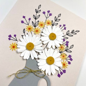 May include: A grey ceramic vase with a gold twine bow holds a bouquet of white daisies with yellow centres and purple and yellow wildflowers.