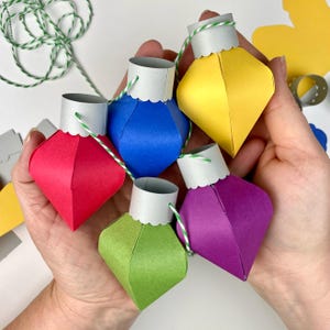 May include: Five paper Christmas ornaments in different colors, strung together with twine. The ornaments are red, blue, yellow, green, and purple.