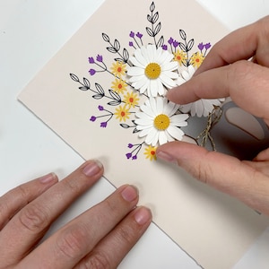 May include: A white card with a hand placing a white paper daisy on top of a bouquet of paper flowers. The bouquet is made of white daisies with yellow centres, yellow flowers with orange centres, and purple flowers. The flowers are arranged in a vase with a grey paper base and a brown twine bow.