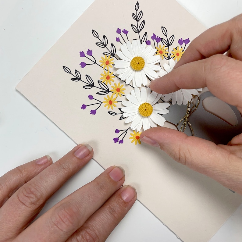 May include: A white card with a hand placing a white paper daisy on top of a bouquet of paper flowers. The bouquet is made of white daisies with yellow centres, yellow flowers with orange centres, and purple flowers. The flowers are arranged in a vase with a grey paper base and a brown twine bow.
