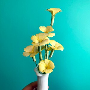 May include: A bouquet of yellow carnations in a white ceramic vase against a turquoise background.