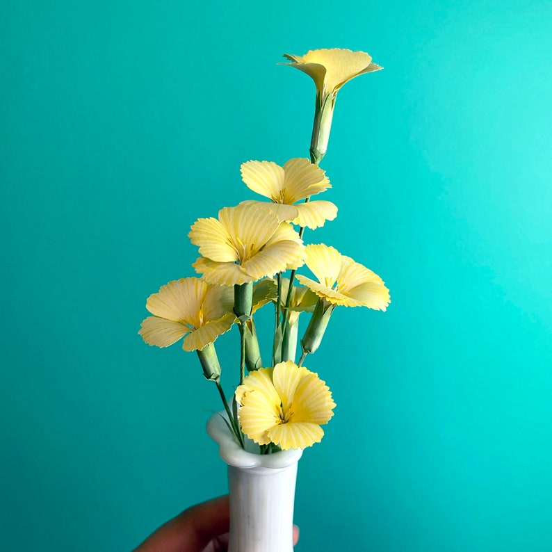 May include: A bouquet of yellow carnations in a white ceramic vase against a turquoise background.