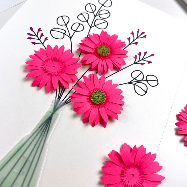 May include: A handmade card featuring a bouquet of pink paper flowers with green centres. The flowers are arranged in a vase and are surrounded by black and white leaves.