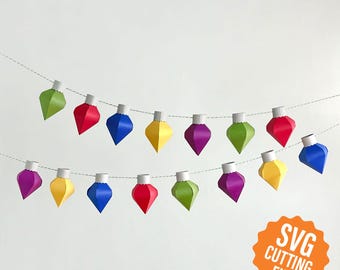 May include: A string of paper Christmas lights with red, green, blue, yellow, and purple ornaments. The lights are strung on a white string and are hanging from a white wall. The text "SVG CUTTING FILE" is in the lower right corner of the image.