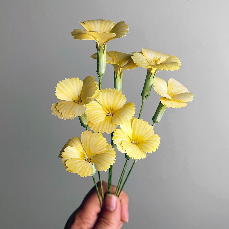 May include: A bouquet of yellow paper flowers with green stems. The flowers are arranged in a fan shape and have a delicate, layered appearance.