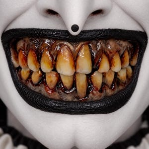 May include: A close-up of a mouth with white skin makeup, thick black lipstick, and a black dot on the philtrum. The teeth appear stained, yellowed, and decayed with dark red, blood-like colouring along the gum line and between the teeth.