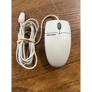 May include: A beige wired computer mouse featuring the Microsoft logo printed in black on the top shell. The device includes two primary buttons, a central grey scroll wheel, and a long white cable ending in a round PS/2 connector plug.
