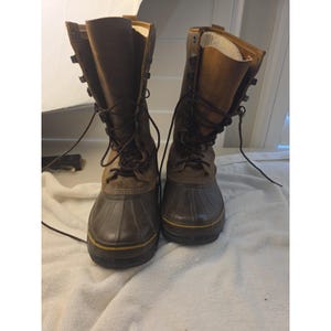 May include: A pair of tall brown leather boots with dark rubber lower shells and thick black soles. The boots feature a lace-up front closure with metal eyelets and hooks, brown laces, and a light-coloured interior lining visible at the top opening.