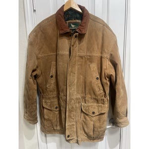 Puede incluir: Una chaqueta de ante color tostado con cuello de cuero marrón, cierre frontal de botones y dos grandes bolsillos con solapa en la cintura. La chaqueta presenta un patrón de diamantes acolchado en el pecho y un forro interior oscuro con etiqueta.