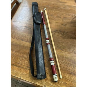 Könnte beinhalten: Ein zweiteiliges Billard-Queue mit einem cremefarbenen Oberteil und einem kastanienbraunen Unterteil, das mit einem silbernen, strukturierten Griffband, weißen Ringen und einem schwarzen Logo versehen ist, inklusive einer schwarzen Tasche.
