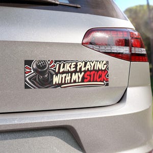 May include: A rectangular bumper sticker featuring a graphic of a black manual transmission gear shift knob with a six-speed pattern. The text reads I LIKE PLAYING WITH MY STICK in bold white and red lettering against a black background.