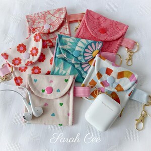 May include: A selection of handmade fabric pouches with floral and geometric designs. Each pouch features a button closure, a pink strap, and a gold-coloured keychain. One pouch contains white earbuds, and another holds a white charging case.