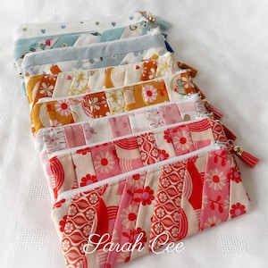 May include: A collection of handmade fabric pencil cases in various floral and patterned designs. Each case features a white zipper and a tassel. The color palette includes pinks, reds, oranges, and blues. The name "Sarah Cee" is printed on the bottom.