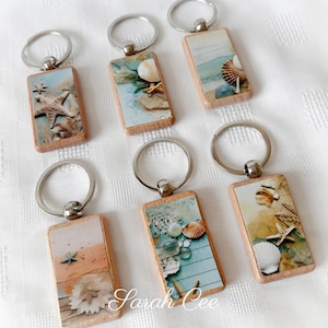 May include: Seven wooden keyrings with beach-themed designs. Each keyring features a rectangular wooden base with a metal ring. The designs include seashells, starfish, and coastal imagery in shades of blue, beige, and white.