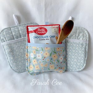 Peut inclure: Un ensemble de maniques et de dessous de plat bleu clair. Une poche à motifs floraux contient un paquet de mélange à biscuits aux pépites de chocolat Betty Crocker et une cuillère en bois. Bordure blanche et boucles.