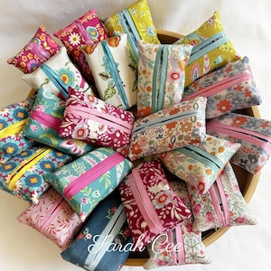 May include: A collection of 15 handmade tissue box covers in various floral patterns and colours, including pink, blue, green, and red. The covers are made of fabric and have a ribbon detail. The covers are arranged in a basket.