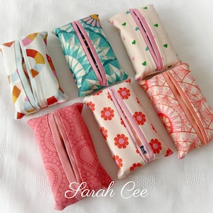 May include: Six fabric tissue box covers with diverse patterns and colours. Designs include geometric shapes, flowers, and hearts. Each cover has a central slit with a pink border for tissue dispensing. The name "Sarah Cee" is printed on the bottom.