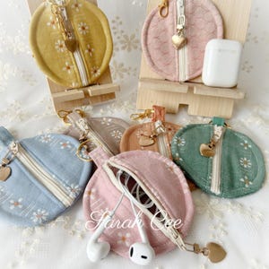 May include: A collection of round, zippered fabric pouches in mustard yellow, dusty pink, light blue, sage green, and beige, each featuring a white daisy pattern and a gold heart-shaped zipper pull. One pouch holds white wired earbuds.