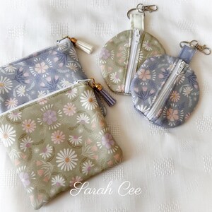 Peut inclure: Quatre pochettes et porte-monnaie à imprimé floral dans les tons de vert et de bleu. Les pochettes sont rectangulaires avec une fermeture éclair et un pompon. Les porte-monnaie sont ronds avec une fermeture éclair et un mousqueton. Le motif floral comprend des fleurs blanches, roses et violettes.