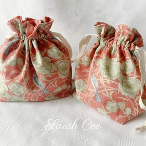 May include: Two drawstring bags with a floral and bird pattern in coral, sage green, and grey. The bags have cream-coloured drawstrings and are gathered at the top. The text "Sarah Cee" is at the bottom of the image.