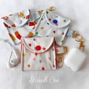 May include: Three small fabric pouches with snap closures, each with a different pattern. One has a colourful geometric design, another has hearts, and the third has stars. Gold-coloured keychains and white earbuds are also visible.