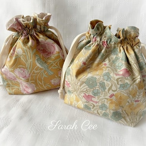 Könnte beinhalten: Zwei Kordelzugbeutel mit Blumen- und Vogelmustern. Eine Tasche hat einen senfgelben Hintergrund mit rosa Blumen und einem blauen Vogel. Die andere Tasche hat einen cremefarbenen Hintergrund mit rosa Vögeln und blauen Blumen. Beide Taschen haben cremefarbene Kordelzüge.