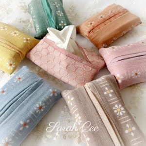 Könnte beinhalten: Eine Sammlung von sieben rechteckigen Taschentuchhaltern aus Stoff in verschiedenen Pastellfarben wie Blau, Gelb, Grün, Rosa und Beige. Jeder Halter hat ein weißes Gänseblümchenmuster und eine mittige Öffnung. Ein rosa Halter enthält ein weißes Taschentuch.