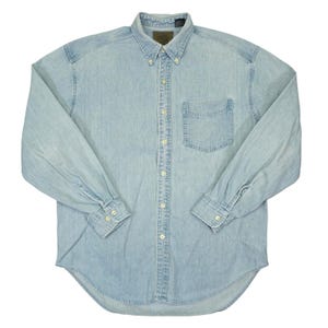 Puede incluir: Una camisa de manga larga de color azul claro con botones, confeccionada en tela de mezclilla. La camisa presenta un cuello puntiagudo con botones, un bolsillo en el pecho en el lado izquierdo y puños con dos botones cada uno. Tiene siete botones visibles.