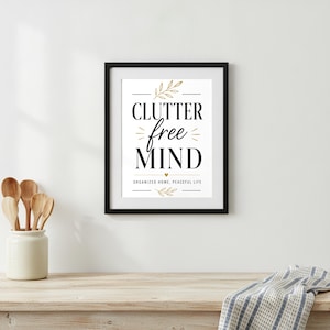 May include: A framed wall art print featuring the black text CLUTTER free MIND with gold leaf accents and the words ORGANIZED HOME, PEACEFUL LIFE printed below. The design includes small gold sprigs and a tiny heart icon on a white background.