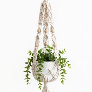 May include: A cream-coloured macrame plant hanger featuring intricate knotted patterns and smooth, round wooden beads. The hanger supports a plain white ceramic pot containing a green artificial plant with small leaves, finished with a tassel.