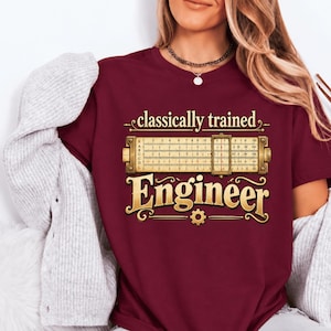 Può includere: Una t-shirt a maniche corte color bordeaux con la grafica di un regolo calcolatore, il testo "classically trained" sopra e la parola "Engineer" sotto in carattere dorato, accentuata da una piccola icona a forma di ingranaggio in basso.