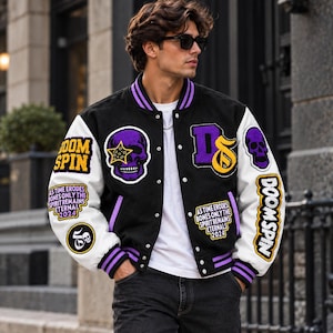 Può includere: Una giacca varsity nera e bianca con toppe ricamate viola e gialle, tra cui teschi, stelle e il testo "DOOM SPIN" e "AS TIME ERODES BONES ONLY THE SPIRIT REMAINS ETERNAL 2024". La giacca presenta una chiusura frontale con bottoni a pressione.