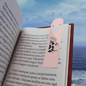 Könnte beinhalten: Ein hellrosa Lesezeichen mit einer schwarz-weißen Kuh-Illustration und dem in Schwarz gedruckten Text MOOMARK. Das Lesezeichen steckt in einem aufgeschlagenen Buch mit lateinischem Text, vor einem unscharfen Küstenhintergrund.