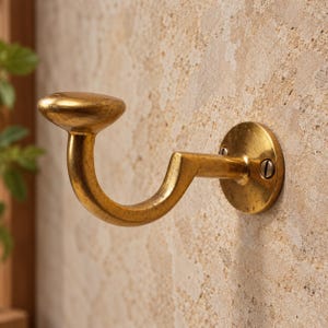 Puede incluir: Un gancho de pared de metal dorado con un brazo curvo y un pomo redondeado y plano en el extremo. El gancho cuenta con una placa de montaje circular fijada a la pared con dos tornillos visibles, mostrando un acabado metálico texturizado y desgastado.