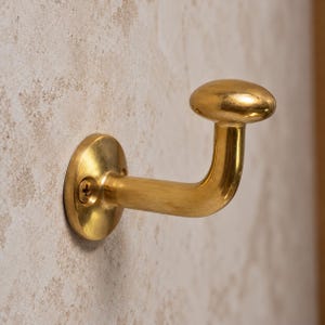 May include: A polished brass wall hook featuring a circular mounting plate with a single visible screw and a curved arm that terminates in a smooth, rounded knob end. The metal surface has a reflective, warm golden tone and a metallic finish.