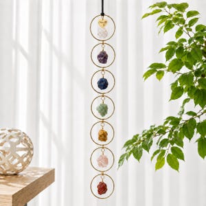 May include: A vertical hanging ornament featuring seven gold-toned metal rings connected in a chain. Each ring holds a raw, unpolished gemstone in various colors, including clear, purple, blue, green, yellow, pink, and red, with a heart charm.