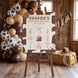 May include: A rectangular milestone sign with a brown and white cow print border, featuring the text Cooper's First Rodeo Milestones, a photo of a child in a party hat, and lists of developmental achievements and favorite songs.