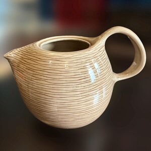 May include: A beige ceramic teapot with a brown striped pattern. The teapot has a rounded body and a curved handle.
