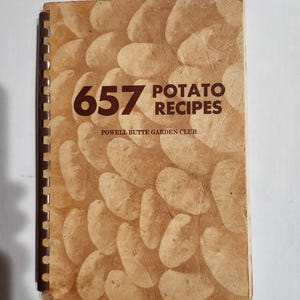 May include: A spiral-bound cookbook with a tan cover featuring a repeating potato pattern. The text on the cover reads 657 POTATO RECIPES and POWELL BUTTE GARDEN CLUB in dark brown lettering. The book has a dark brown plastic binding spine.