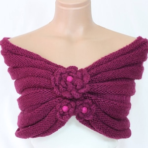 May include: A knitted burgundy shawl with a floral design. The shawl features a ribbed texture and is adorned with two flower accents and three pink glittery embellishments. The garment is displayed on a mannequin.
