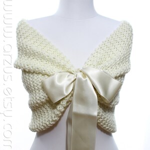 May include: A white knitted shawl with a large ivory satin bow. The shawl is draped over a white mannequin.